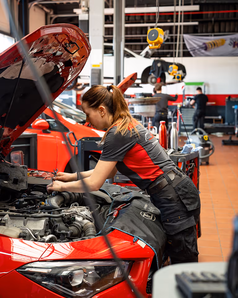 Mechanikerin arbeitet in der Werkstatt am Motor eines roten Autos mit geöffneter Motorhaube