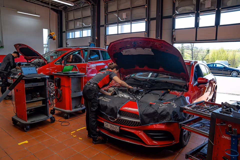 Zwei Mechaniker arbeiten in der Werkstatt an Fahrzeugen, vorne ein roter Mercedes mit geöffneter Motorhaube und Schutzabdeckung