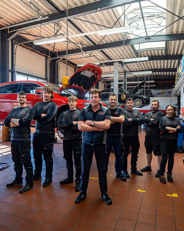 Das Werkstatt-Team steht mit verschränkten Armen in der Werkstatt, im Hintergrund ein rotes Auto mit geöffneter Motorhaube auf der Hebebühne