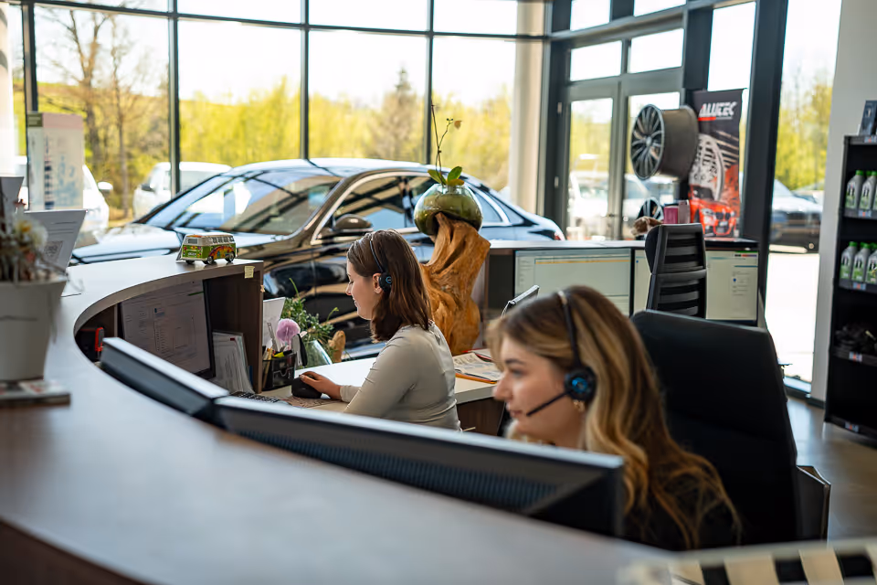 Zwei Mitarbeiterinnen mit Headset am Empfangstresen im Autohaus Schmid, dahinter ein Fahrzeug im verglasten Ausstellungsraum